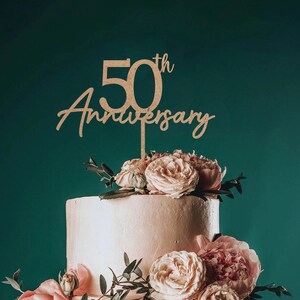 May include: Wooden cake topper with the text "50th Anniversary" in a cursive font. The topper is placed on a two-tiered cake decorated with pink roses and greenery.