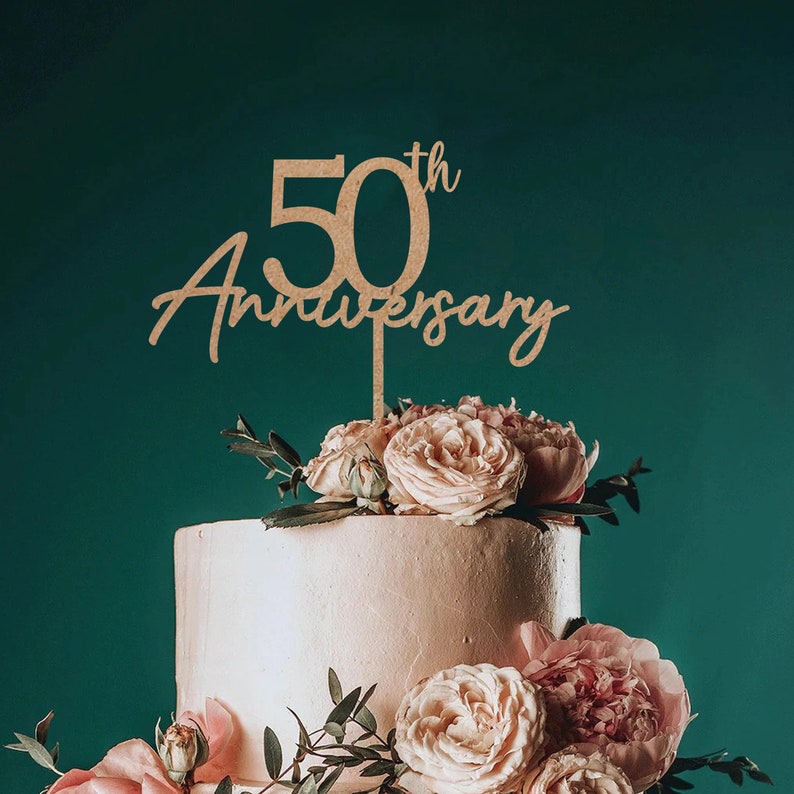 May include: Wooden cake topper with the text "50th Anniversary" in a cursive font. The topper is placed on a two-tiered cake decorated with pink roses and greenery.