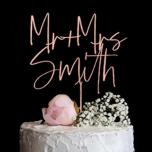 Puede incluir: Una tarta de boda con glaseado blanco, coronada con un adorno para tarta "Mr + Mrs Smith" en oro rosa. La tarta est&aacute; decorada con una peon&iacute;a rosa y flores blancas sobre un fondo negro.
