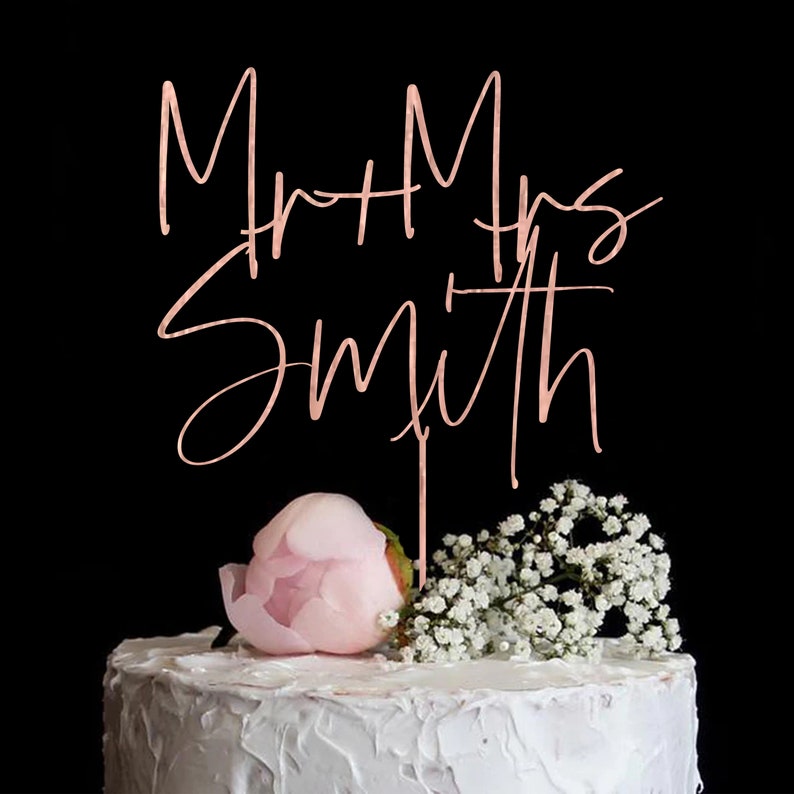 Puede incluir: Una tarta de boda con glaseado blanco, coronada con un adorno para tarta "Mr + Mrs Smith" en oro rosa. La tarta est&aacute; decorada con una peon&iacute;a rosa y flores blancas sobre un fondo negro.