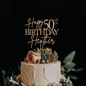 Pu&ograve; includere: Un cake topper in legno con la scritta "Happy 50th Birthday Heather" in elegante corsivo. Il topper &egrave; posto su una torta glassata bianca decorata con fiori e verde, su uno sfondo scuro.