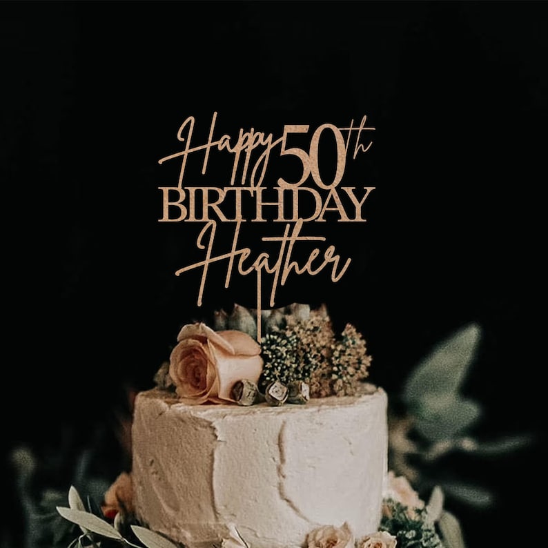 Pu&ograve; includere: Un cake topper in legno con la scritta "Happy 50th Birthday Heather" in elegante corsivo. Il topper &egrave; posto su una torta glassata bianca decorata con fiori e verde, su uno sfondo scuro.