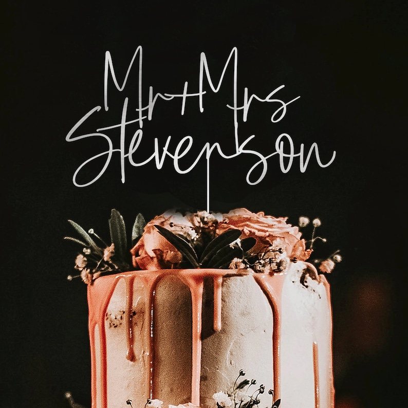 Puede incluir: Una tarta de boda con goteo de glaseado color coral y decoraciones florales. Un adorno plateado para tarta muestra "Mr + Mrs Stevenson" en escritura cursiva sobre un fondo oscuro.