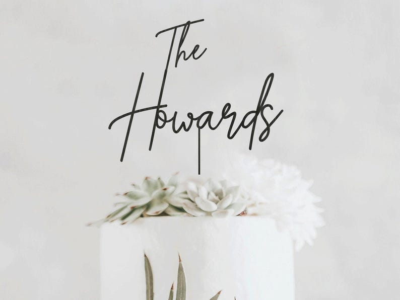 May include: A black script cake topper that reads "The Howards" is placed on top of a white cake decorated with succulents and white flowers. The cake is set against a soft, neutral background.