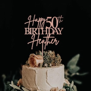 Pu&ograve; includere: Un cake topper color oro rosa con la scritta "Happy 50th Birthday Heather" &egrave; posto sopra una torta glassata bianca decorata con fiori e verde. La torta &egrave; ambientata su uno sfondo scuro.