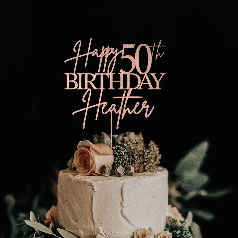 Pu&ograve; includere: Un cake topper color oro rosa con la scritta "Happy 50th Birthday Heather" &egrave; posto sopra una torta glassata bianca decorata con fiori e verde. La torta &egrave; ambientata su uno sfondo scuro.