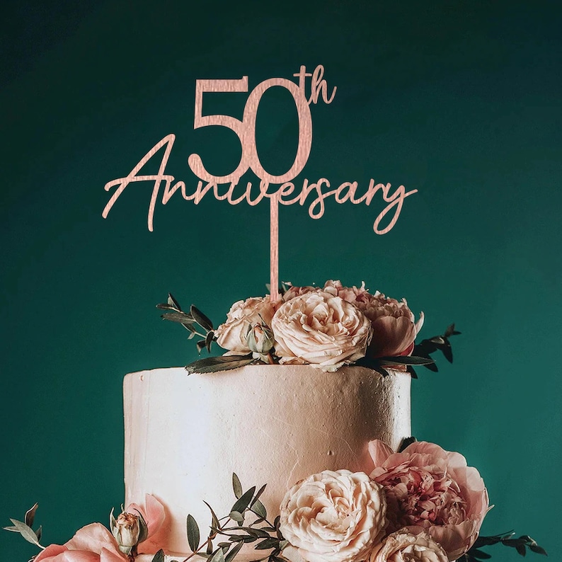 May include: A two-tiered white cake decorated with pink roses and greenery. A wooden cake topper with the text "50th Anniversary" is placed on top of the cake.
