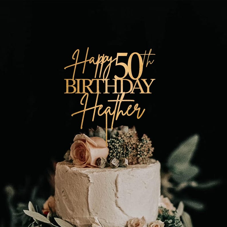 Pu&ograve; includere: Un cake topper dorato con la scritta "Happy 50th Birthday Heather" in un elegante carattere. Il topper &egrave; posto su una torta glassata bianca decorata con fiori e verde, su uno sfondo scuro.