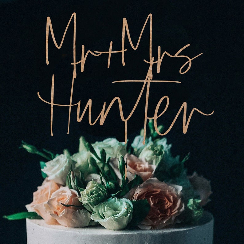 Puede incluir: Una tarta blanca con un ramo de rosas en tonos rosa, melocot&oacute;n y blanco. La tarta est&aacute; coronada con las palabras "Mr + Mrs Hunter" en una fuente dorada sobre un fondo oscuro.