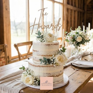 May include: A two-tiered wedding cake with white frosting and floral decorations. The cake is topped with a wooden sign that reads "The Howards." The cake is on a wooden table with a floral arrangement and candles in the background.