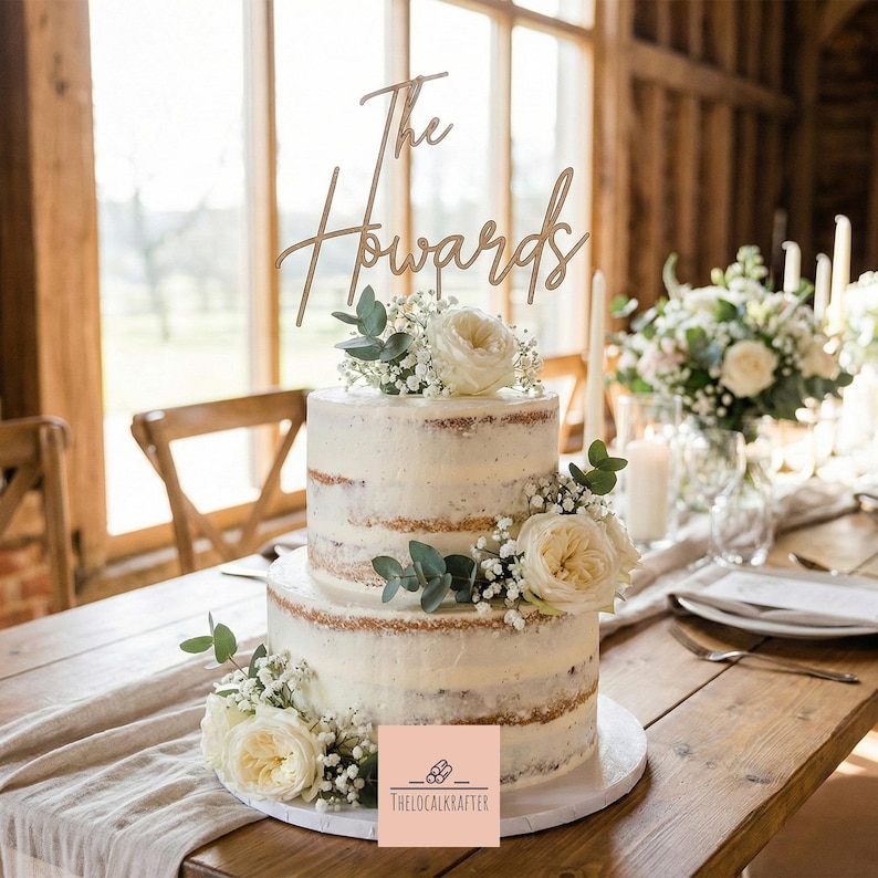 May include: A two-tiered wedding cake with white frosting and floral decorations. The cake is topped with a wooden sign that reads "The Howards." The cake is on a wooden table with a floral arrangement and candles in the background.