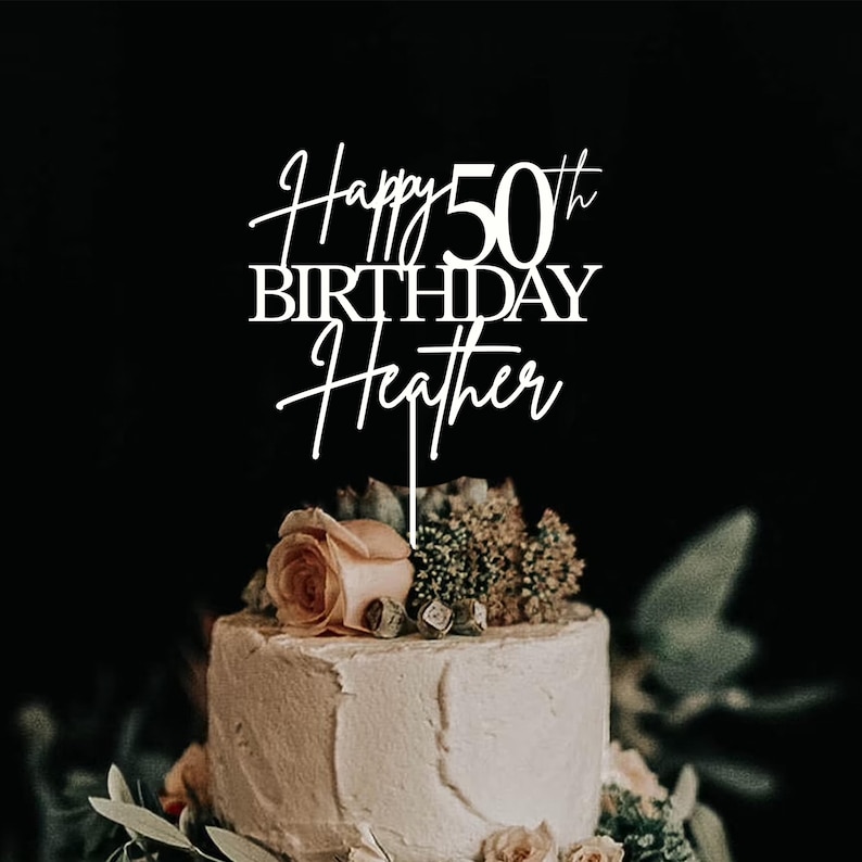 Pu&ograve; includere: Una torta bianca decorata con una composizione floreale e un cake topper con la scritta "Happy 50th Birthday Heather". La torta &egrave; adornata con fiori e verde. Lo sfondo &egrave; scuro.