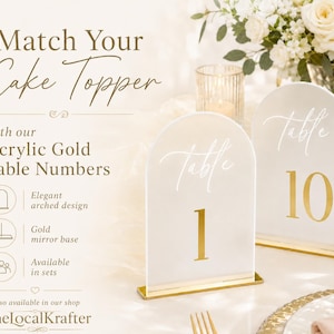 May include: Two acrylic gold table numbers with arched design. The numbers are white with gold accents and bases. The text on the signs reads "Table 1" and "Table 10". The signs are displayed on a table with flowers and a candle.