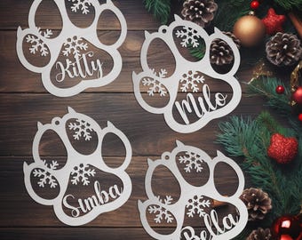 May include: Four white paw print ornaments with snowflake designs and personalized names: Kitty, Milo, Simba, and Bella. The ornaments are hanging on a brown wooden surface with Christmas greenery and ornaments in the background.
