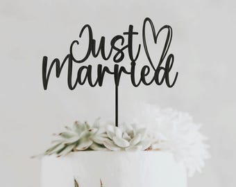 Puede incluir: Un adorno negro para pastel con las palabras "Just Married" en fuente cursiva, con un corazón reemplazando la letra "i". El adorno está colocado sobre un pastel blanco decorado con flores blancas y suculentas.