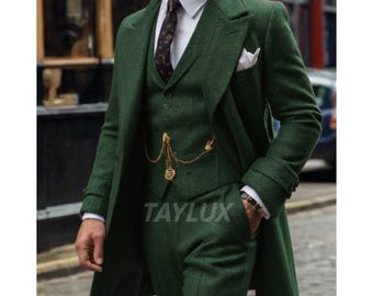 May include: A dark green three-piece suit with a long coat. The suit includes a vest, pants, and a coat with a pocket square. A gold chain is visible on the vest. The text "TAYLUX" is printed on the pants.