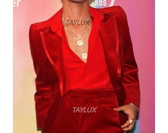 Può includere: Un completo in velluto rosso, composto da una giacca e pantaloni abbinati. La giacca ha un classico revers a lancia e una chiusura a due bottoni. L'abito è abbinato a una camicia rossa abbottonata e collane d'oro. Il marchio "TAYLUX" è visibile.