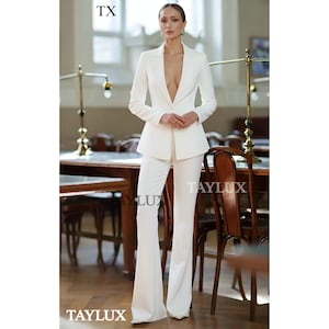 May include: A white suit comprising a blazer and flared trousers. The blazer features a deep V-neck. The image includes the text "TX" and "TAYLUX". The suit is worn in a room with wooden tables and chairs.