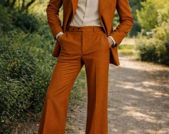 Pode incluir: Um fato cor de ferrugem com um blazer a condizer e calças de corte largo. O fato é usado sobre uma camisa creme. A imagem é tirada ao ar livre com um caminho e vegetação no fundo. A marca "TAYLUX" é visível.