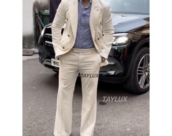 May include: A cream-colored suit with a double-breasted blazer and wide-leg pants. The blazer has gold buttons and a textured design. The person is wearing a light blue shirt and tan shoes. The word "TAYLUX" is printed on the pants.