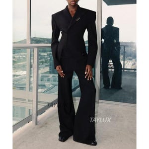 Puede incluir: Un traje negro con una americana entallada con hombros estructurados y pantalones de pernera ancha. La americana tiene un cierre decorativo. Los pantalones son acampanados. La palabra "TAYLUX" está impresa en los pantalones.