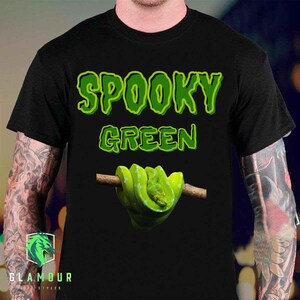 Peut inclure: Un t-shirt noir avec le texte SPOOKY GREEN écrit dans une police de caractères vert citron dégoulinante. Sous le texte se trouve un graphique représentant un serpent vert vif enroulé autour d'une branche d'arbre brune sur le torse du vêtement.