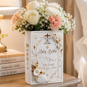 Könnte beinhalten: Eine weiße Keramikvase in Buchform mit der Aufschrift "Holy Bible" und einem religiösen Zitat in Gold. Die Vase ist mit floralen Details verziert und enthält einen Strauß aus weißen Rosen, rosa Nelken und Schleierkraut. Die Vase steht auf einer Holzoberfläche.