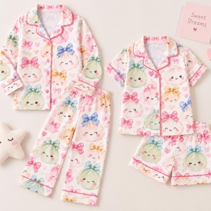 May include: Two sets of pajamas featuring a white fabric with a repeating pattern of smiling dumplings wearing colorful bows and small pink hearts. One set includes a long-sleeve button-down shirt and long pants, while the other has a short-sleeve shirt and shorts.