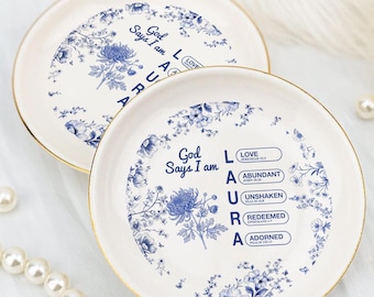 May include: Two white decorative plates with blue floral designs and gold rims. One plate has the text "God Says I am LAURA" with associated words. The other plate has the text "God Says I am ARCA". Pearls and a gold ring are also in the image.