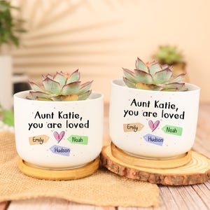 May include: Two white ceramic planters with succulents, each inscribed with "Aunt Katie, you are loved." The planters have wooden saucers. The text includes names: Emily, Noah, and Hudson. A heart graphic is also included.