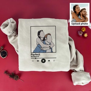 May include: A beige sweatshirt with a graphic of a couple hugging. The graphic is in a black frame with the text "Perfect" and "Ed Sheeran" below it. There are music player controls below the graphic.