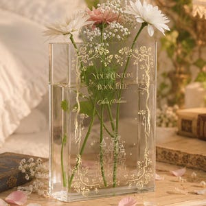 May include: A clear glass vase with a rectangular shape, filled with water and flowers. The vase has a gold decorative border with the text "YOUR CUSTOM BOOK TITLE" and "Olivia Williams". The flowers include white and pink daisies.
