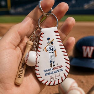 Könnte beinhalten: Ein Baseball-Schlüsselanhänger mit einem weißen Lederanhänger mit einer Baseballspieler-Illustration und dem Text "GOD HIT A HOMERUN WHEN HE MADE YOU My Dad." Enthält einen kleinen Baseballschläger und einen Baseball-Charm.