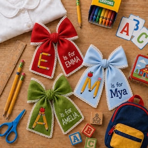 Op de afbeelding: Drie stoffen haarstrikken in rood, lichtblauw en groen, elk met witte kanten rand, een geborduurde letter van potloden en de tekst 'is for' gevolgd door een naam: EMMA, Mya of AMELIA. De strikken liggen tussen diverse schoolspullen.