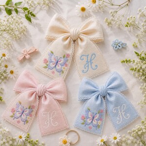 Può includere: Tre fiocchi per capelli in tessuto color crema, rosa e azzurro, ciascuno con bordo in pizzo, una farfalla ricamata e la lettera H. Sono inclusi anche una piccola clip per capelli a forma di farfalla azzurra e un accessorio a fiore bianco.