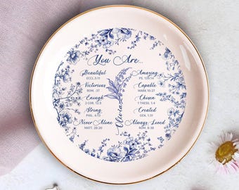 May include: A round, off-white decorative plate with a gold rim. The plate features a blue floral design around the edge and inspirational text in the center, including the name "Olivia".
