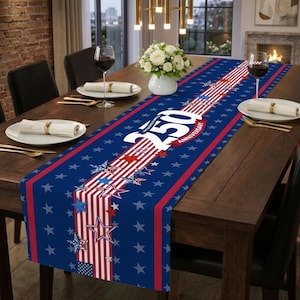 Puede incluir: Camino de mesa azul con un diseño patriótico con rayas rojas y blancas, estrellas y el texto "250 ANIVERSARIO". El camino de mesa está colocado sobre una mesa de madera, con cubiertos y copas de vino visibles.