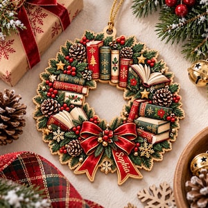 May include: A circular wooden Christmas ornament featuring a wreath design with miniature books, pinecones, green pine needles, red berries, and gold stars. A large red ribbon is at the bottom with the name Sophia printed in white script.