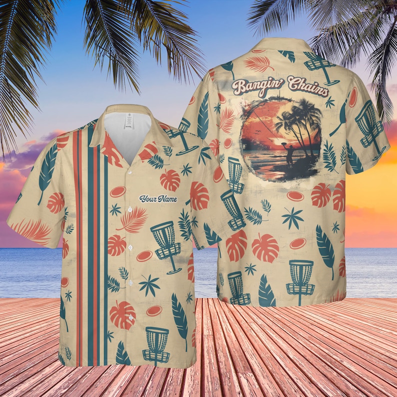 May include: A beige short-sleeved button-up shirt with a tropical print featuring palm trees, leaves, and disc golf discs. The shirt has a pocket on the left side with the text "Your Name" printed on it. The back of the shirt has a sunset scene with the text "Bangin' Chains".