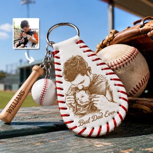 Könnte beinhalten: Ein Baseball-Schlüsselanhänger mit weißem Lederdesign, roten Nähten und einem Metallring. Der Schlüsselanhänger zeigt ein eingraviertes Bild eines Vaters und Kindes mit dem Text "Best Dad Ever". Ein kleiner Holzschläger mit "WILLIAMS" ist befestigt.