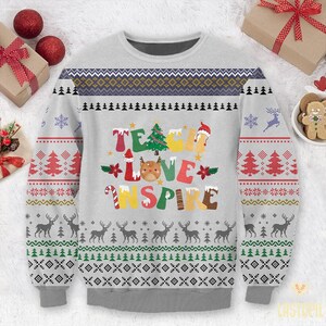Puede incluir: Un suéter navideño gris con un diseño festivo que presenta las palabras "Teach Love Inspire" en letras de colores. El suéter tiene un patrón de copo de nieve rojo y verde en las mangas y un patrón de copo de nieve blanco y gris en el cuerpo.