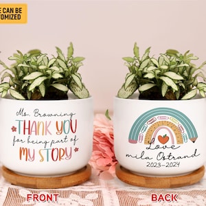 May include: White ceramic planter with a green plant. The front of the planter says "Ms. Browning THANK YOU for being part of MY STORY*" and the back says "Love mila Ostrand 2023-2024" with a rainbow design featuring school supplies.