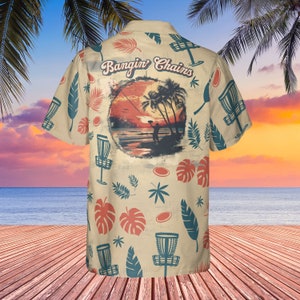 May include: A beige short-sleeved button-up shirt with a tropical print. The shirt features a sunset scene with a person throwing a disc, palm trees, and the text "Bangin' Chains".