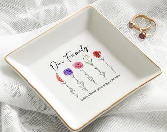 May include: A white ceramic ring dish with gold trim, featuring the text "Our Family" and floral illustrations. The dish also has the phrase "Creating a forever garden of love in our home." Two gold rings with red gemstones are on the dish.