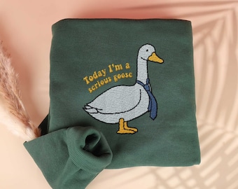 May include: A dark green sweatshirt with a white goose wearing a blue tie embroidered on the front. The text "Today I'm a serious goose" is embroidered in yellow above the goose.