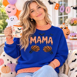 Könnte beinhalten: Ein königsblaues Sweatshirt mit Stehkragen und Ärmelbündchen in orange-weißem Vichy-Karo. Auf der Vorderseite steht das Wort MAMA in orangefarbenem Vichy-Karo mit Tropfeffekt, zwei Skeletthände halten Kürbisse und kleine schwarze Fledermäuse.