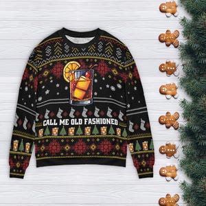 Op de afbeelding: Zwarte kersttrui met een cocktailafbeelding en de tekst "CALL ME OLD FASHIONED". De trui heeft een feestelijk patroon met sneeuwvlokken, bomen en cadeauverpakkingen. Peperkoekkoekjes en dennentakken zijn rond de trui gerangschikt.