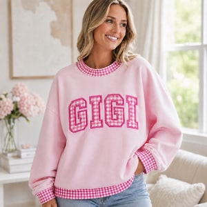 Könnte beinhalten: Ein hellrosa Sweatshirt mit Rundhalsausschnitt und dem Wort GIGI in großen, rosa-weiß karierten Blockbuchstaben auf der Brust. Das Kleidungsstück hat passende karierte Abschlüsse am gerippten Halsausschnitt, an den Ärmelbündchen und am Saum.