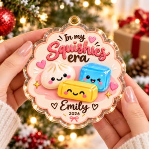 Könnte beinhalten: Ein gewelltes Acryl-Ornament mit dem Text In my Squishies era, Emily und 2026. Das Design zeigt ein weißes Knödel, einen blauen Eiswürfel und einen gelben rechteckigen Block, alle mit lächelnden Gesichtern, rosa Wangen und kleinen Herzen.