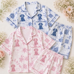 Könnte beinhalten: Zwei Sets aus kurzärmeligen, geknöpften Pyjama-Oberteilen und passenden Shorts, eines in Blau-Weiß und eines in Rosa-Weiß. Beide zeigen ein wiederkehrendes Muster aus Dackeln, Blumenmotiven und Briefmarkendesigns mit dem Text Carte Postale.